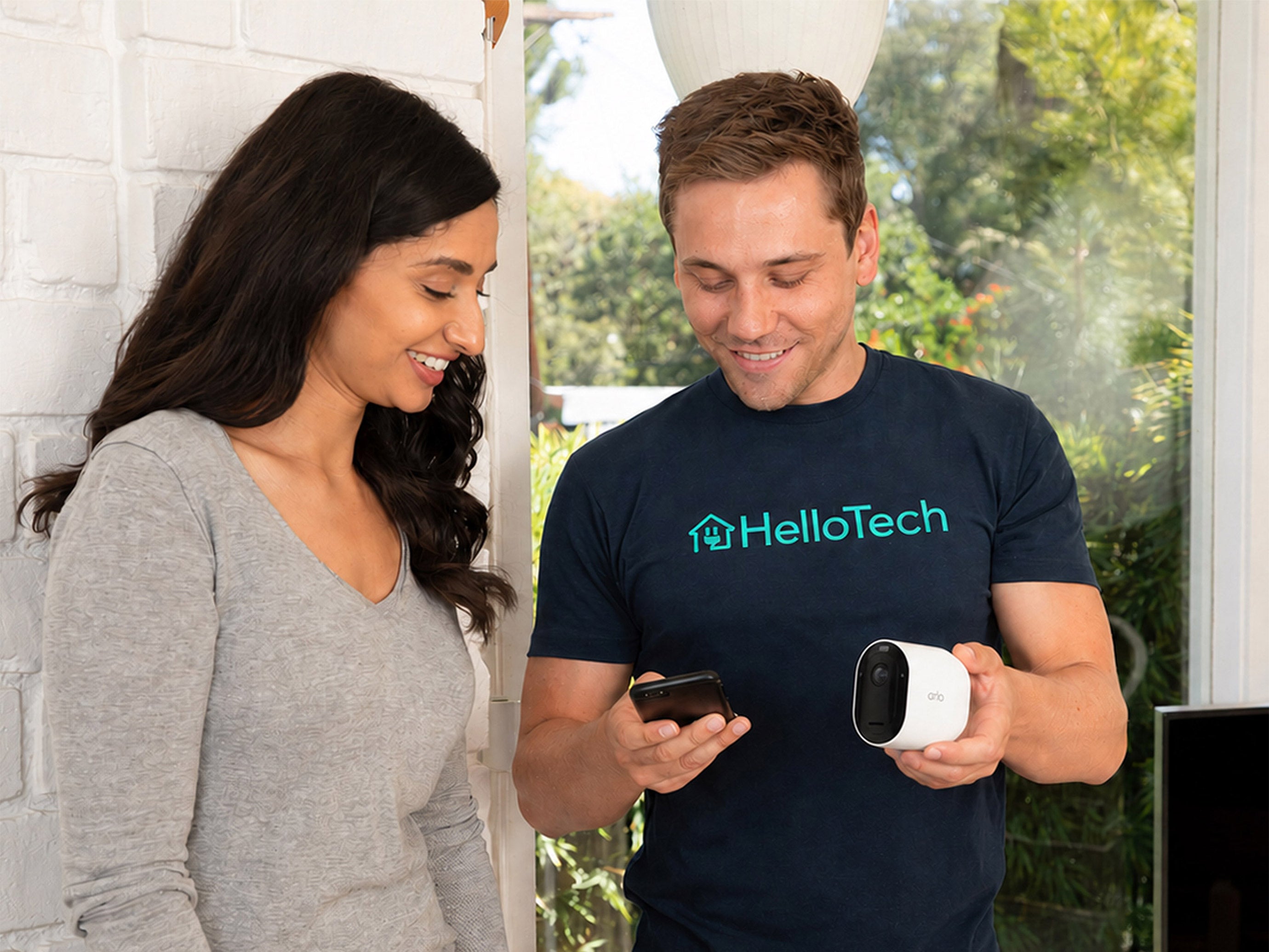 A man wearing a navy HelloTech t-shirt holds a Arlo Pro Security Camera (6th Gen) while looking at a phone, standing next to a woman in a gray long-sleeve top in a bright indoor setting.